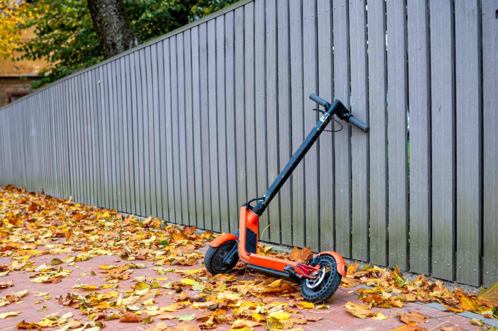 Embedos_electric-scooter-edge-sidewalk-covered-with-autumn-leaves-by-wooden-fence